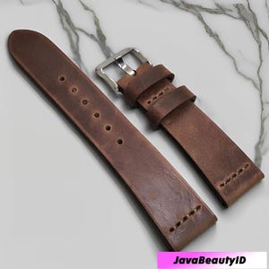 May include: Two-piece brown leather watch strap with a silver buckle. The strap has adjustment holes and is designed for a watch. The leather has a textured appearance.
