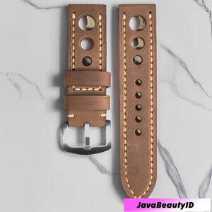 Op de afbeelding: Twee bruine leren horlogebandjes met crèmekleurige stiksels en een zilveren gesp. Eén bandje heeft drie grote gaten en de andere zes kleinere gaten. De bandjes worden gepresenteerd op een wit marmeren oppervlak.
