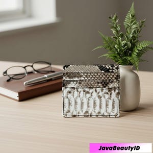 Pode incluir: Porta-cartões com padrão de pele de cobra em tons de branco, cinza e preto, com várias ranhuras para cartões. Está sobre uma superfície de madeira clara, ao lado de um pequeno vaso com vegetação, um caderno, óculos e uma caneta.