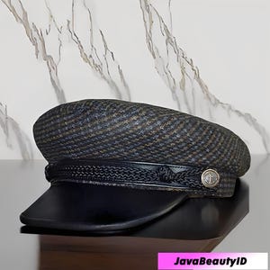 Peut inclure: Casquette gavroche en tweed foncé avec une visière en cuir noir. La casquette est ornée d'une bande décorative et d'un bouton argenté avec un emblème. Le texte "JavaBeautyID" est visible en bas.
