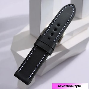 Puede incluir: Correa de reloj de cuero negro con costuras blancas. La correa tiene forma rectangular con bordes redondeados y una hebilla. La correa tiene tres agujeros para ajustar y una presilla para asegurar el exceso de longitud.