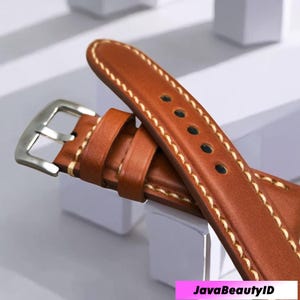 May include: Close-up of a brown leather watch strap with cream stitching. The strap has a silver buckle and adjustment holes. The watch band is a rich, warm brown colour.