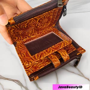 May include: Open, brown leather wallet with a floral embossed pattern. The wallet features a clear ID window, card slots, and a zip closure. The interior is lined with a darker brown fabric. The wallet is open, showing its contents.