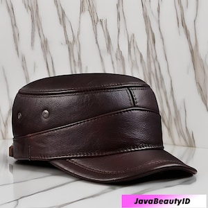 May include: A dark brown leather cap with a short peak. The cap has a band that wraps around the head, secured with two metal rivets. The cap has a structured design and a slightly curved peak. The background is a marble pattern.