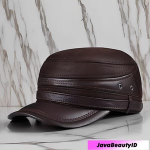 May include: A brown leather cap with a short brim. The cap has a structured design with horizontal stitched bands and two small metal eyelets on the side. The cap is a dark brown color.