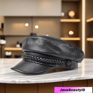 Puede incluir: Gorra de cuero negra con una banda trenzada. La gorra tiene una visera corta y un botón en el lateral. El sombrero está sobre una superficie de mármol. El texto "JavaBeautyID" está en la parte inferior.
