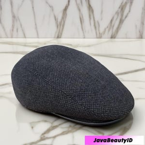 Puede incluir: Gorra plana gris oscuro con tejido texturizado. El sombrero tiene una corona redondeada y una pequeña visera curvada. La gorra probablemente está hecha de un material tejido, y la visera parece ser de cuero o similar.