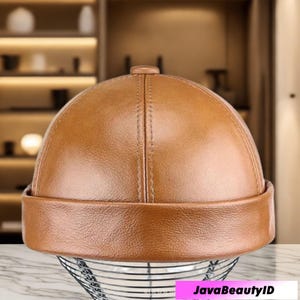 May include: A brown leather cap with a rounded crown and a short, folded brim. The cap has a vertical seam down the center and is displayed on a wire mesh stand. The cap is a warm, medium brown color.