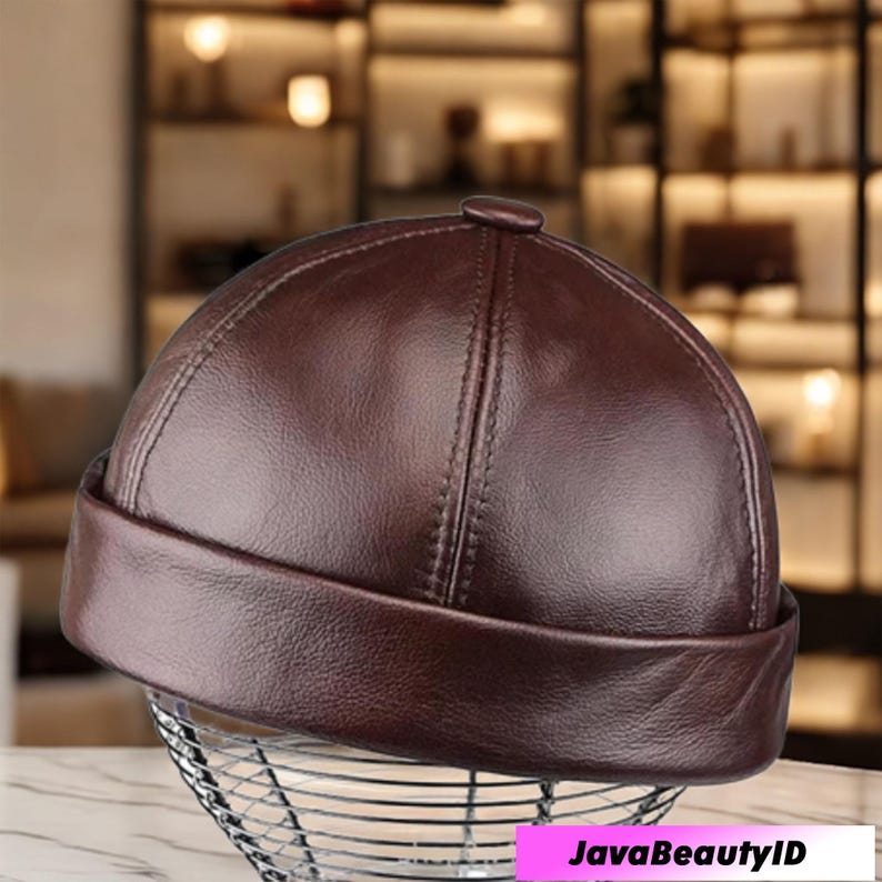 May include: A dark brown leather cap with a rounded crown and a short, turned-up brim. The cap has a stitched seam running down the center and a small button on top. The cap is displayed on a wire mesh stand.