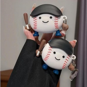 May include: Two baseball-themed accessories, each with a smiling face, red stitching, and a black cap. They have brown arms holding a small bat and blue feet. The accessories are white with a baseball design.