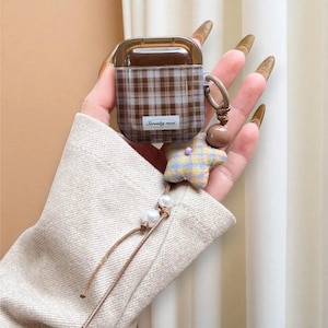 May include: Brown plaid AirPod case with a clear lid, a star-shaped charm, and a beaded lanyard. The case has the text "seventy noon" on the front. The case is held by a person.