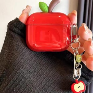 May include: A bright red apple-shaped case for wireless earbuds, with a green leaf and brown stem. A silver-coloured keychain with a red apple charm and green beads is attached to the case.