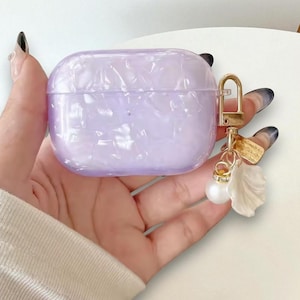 Könnte beinhalten: Ein lavendelfarbenes AirPod-Case mit Perlmutt-Finish. Das Case hat einen goldfarbenen Verschluss und einen Anhänger mit einer Perle, einem Blatt und einem kleinen Anhänger. Das Case wird in einer Hand gehalten.