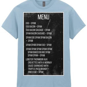 Puede incluir: Camiseta azul claro con un diseño de menú en blanco y negro. El menú enumera varios platos a base de Spam, incluyendo Huevo + Spam, Huevo Bacon + Spam y Langosta Thermidor Aux Crevettes.