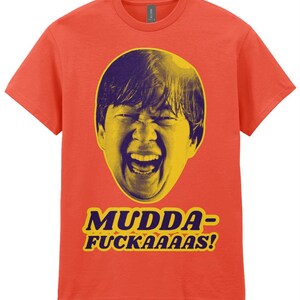 May include: Orange t-shirt featuring a yellow and purple graphic of a person's face with an open mouth. The text "MUDDA-FUCKAAS!" is printed in yellow and purple below the graphic.