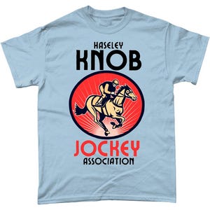 May include: Pale blue t-shirt with a circular design of a jockey on a horse, set against a red background. The text "HASELEY KNOB" is above the graphic, and "JOCKEY ASSOCIATION" is below the image.