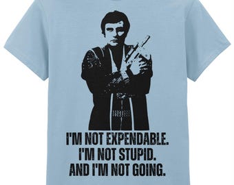 Op de afbeelding: Lichtblauw t-shirt met een zwart silhouet van een persoon die een wapen vasthoudt. De tekst eronder luidt: "I'M NOT EXPENDABLE. I'M NOT STUPID. AND I'M NOT GOING."