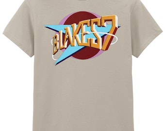 Op de afbeelding: Een beige t-shirt met de tekst "BLAKES 7" in een retro lettertype. Het ontwerp bevat een blauwe bliksemschicht, een rode cirkel en een roze omtrek. Het t-shirt is gemaakt van een zacht materiaal en heeft korte mouwen.
