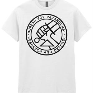 May include: White t-shirt featuring a black circular graphic with the text "BUREAU FOR PARANORMAL RESEARCH AND DEFENSE". The graphic includes a stylized fist and geometric shapes. The shirt is made of a soft material.
