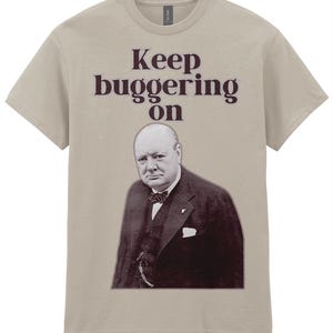 Può includere: Una maglietta beige con un'immagine in bianco e nero di Winston Churchill. Il testo "Keep buggering on" è stampato sopra l'immagine in un colore bordeaux scuro. La maglietta ha un girocollo e maniche corte.