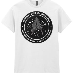 Op de afbeelding: Een wit T-shirt met korte mouwen en een rond zwart embleem op de borst. Het embleem toont het delta-insigne van Starfleet Command, omringd door sterren en de tekst STARFLEET COMMAND UNITED FEDERATION OF PLANETS in het wit.