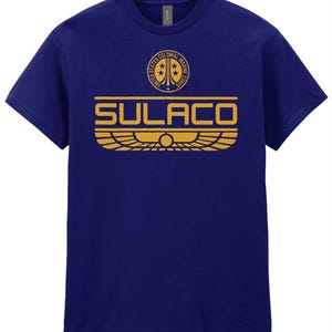 Könnte beinhalten: Ein dunkelblaues Kurzarm-T-Shirt mit einer goldenen Grafik auf der Brust. Das Design zeigt ein kreisförmiges United States Colonial Marine Corps Emblem über dem Wort SULACO, das über einem stilisierten geflügelten Emblem mit einem Mittelkreis liegt.