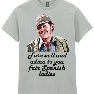 Puede incluir: Camiseta gris claro con un hombre sonriente con gorra y chaqueta. El texto "Farewell and adieu to you fair Spanish ladies" está impreso en escritura negra debajo de la imagen. La camiseta es de material suave.