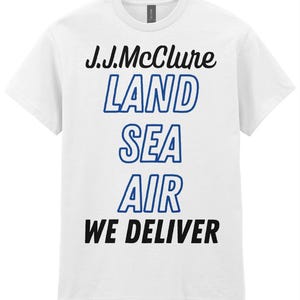 May include: White t-shirt featuring the text "J.J.McClure LAND SEA AIR WE DELIVER" in black and blue lettering. The shirt has a crew neck.