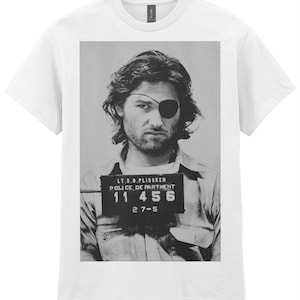 Peut inclure: Un t-shirt blanc à manches courtes avec une impression graphique en noir et blanc représentant un portrait judiciaire. L'image montre une personne aux cheveux longs avec un cache-œil tenant un panneau indiquant LT.S.D.PLISSKEN POLICE DEPARTMENT 11 456 2 7-5.