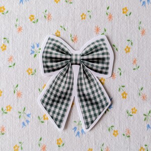 Op de afbeelding: Een groen en wit geruit strikje met een witte rand. De strik staat tegen een witte achtergrond met een bloemenpatroon van gele, oranje en blauwe bloemen.