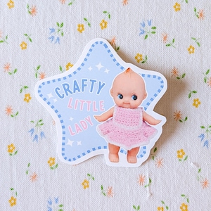 May include: A sticker featuring a baby doll in a pink dress, with the text "Crafty Little Lady" on a light blue star-shaped background. The sticker is set against a floral patterned fabric.