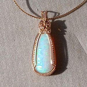 May include: A teardrop-shaped pendant necklace featuring a vibrant opal stone. The opal displays iridescent colors of blue, green, and white, set in a gold-colored filigree frame. The necklace has a delicate gold chain.