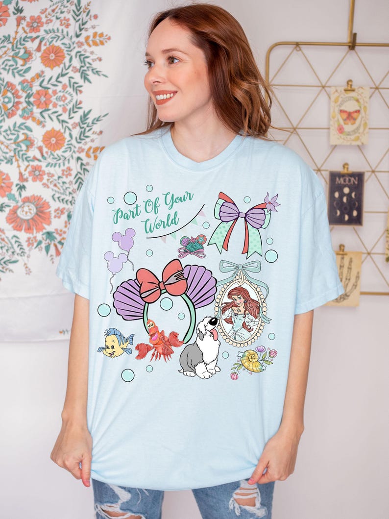 May include: A light blue t-shirt featuring a graphic print with the text Part Of Your World, a mermaid character in an oval frame, a crab, a fish, a dog, a large bow, and mouse-ear headbands with purple seashell designs and bubbles.