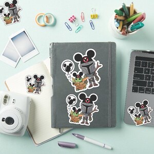 May include: Die-cut vinyl stickers featuring a cartoon Mandalorian and Grogu, both wearing Mickey Mouse ear hats. The Mandalorian holds a Mickey-shaped balloon. The stickers are displayed on a gray notebook, a laptop, and a white notebook, showcasing the character design with gray, green, and brown colors.