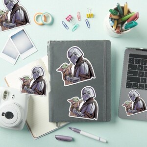 May include: A collection of stickers featuring a character in silver armor holding a small green creature. The stickers are in various sizes and are placed on a gray notebook, a laptop, and a white instant camera. Other office supplies are also visible.