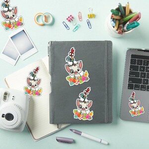 May include: A collection of colorful stickers featuring a cartoon pig character with a rooster on its head, surrounded by flowers. The stickers are placed on a gray notebook, a laptop, and a white camera, with stationery items scattered around.