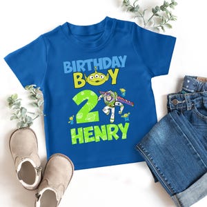 May include: A blue t-shirt with a graphic that reads "BIRTHDAY BOY" and the name "HENRY". The graphic includes a green alien and a cartoon of Buzz Lightyear. A pair of jeans and shoes are next to the shirt.