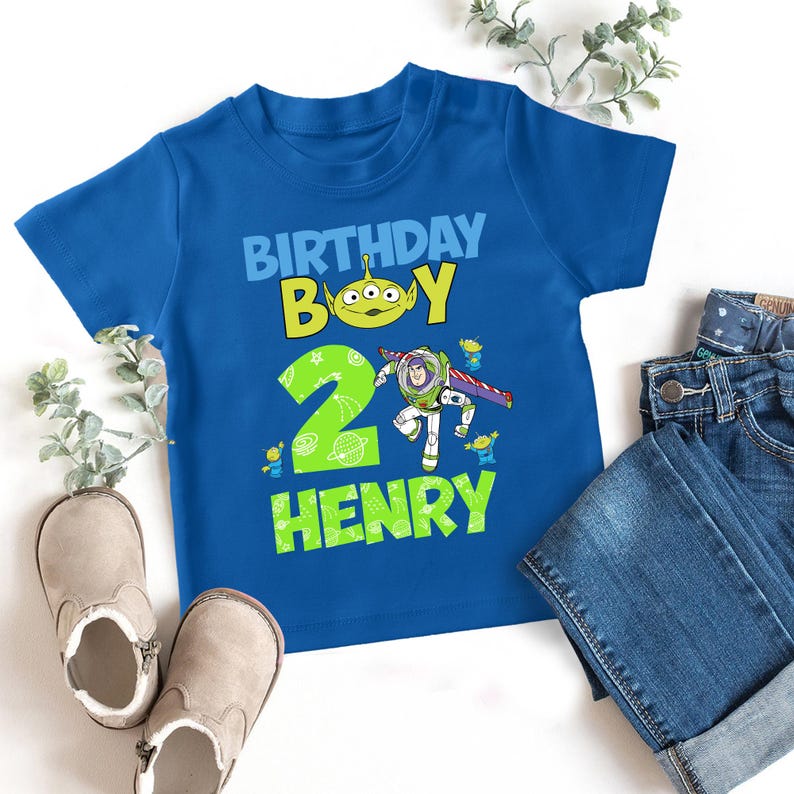 May include: A blue t-shirt with a graphic that reads "BIRTHDAY BOY" and the name "HENRY". The graphic includes a green alien and a cartoon of Buzz Lightyear. A pair of jeans and shoes are next to the shirt.
