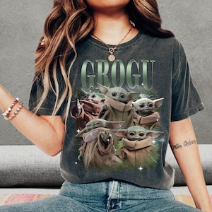 May include: A charcoal gray short-sleeve t-shirt featuring a graphic print of multiple Grogu characters in tan robes. The text GROGU is printed in large green letters above the characters, with green lightning accents surrounding the figures.