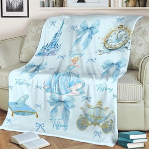 K&ouml;nnte beinhalten: Hellblaue Decke mit Illustrationen einer Prinzessin, eines Schlosses, einer Kutsche, einer Uhr und Schleifen. Der Name "Tiffany" ist in Schreibschrift auf die Decke geschrieben. Die Decke ist &uuml;ber ein beiges Sofa drapiert.