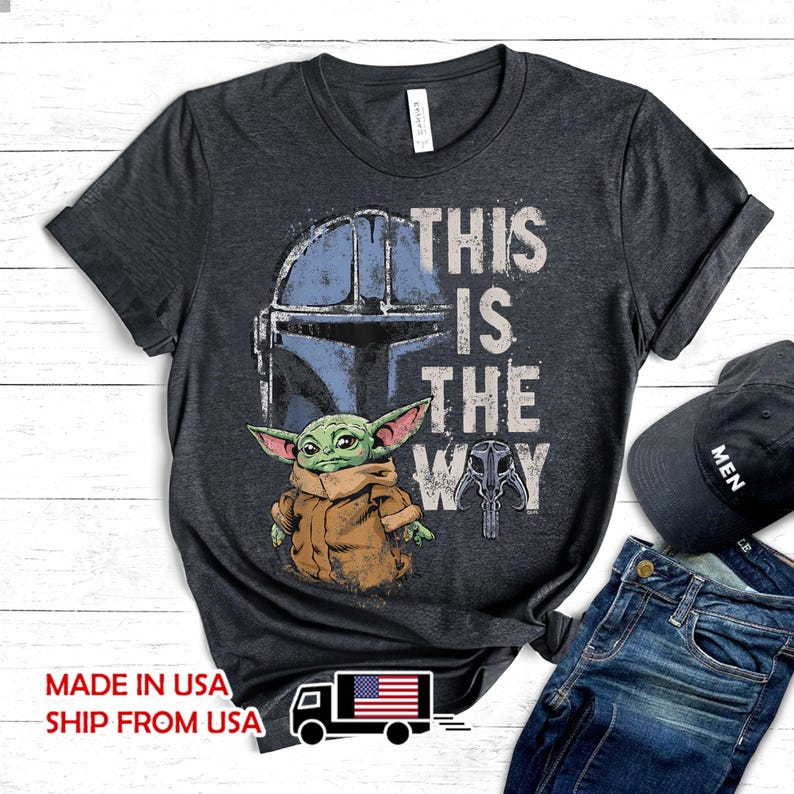 May include: A dark heather gray short-sleeve t-shirt featuring a graphic of a blue Mandalorian helmet and a green creature in a tan robe. The text reads THIS IS THE WAY. A small skull icon is included. The shirt is paired with blue jeans and a black cap.