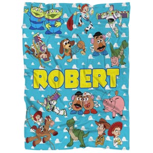 K&ouml;nnte beinhalten: Eine blaue Decke mit Figuren aus dem Film Toy Story. Die Decke hat einen hellblauen Hintergrund mit wei&szlig;en Wolken und dem Namen "ROBERT" in Gelb. Verschiedene Zeichentrickfiguren sind abgebildet.