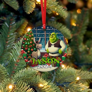 Pode incluir: Enfeite de Natal redondo com Shrek e Burro do filme Shrek, com uma árvore de Natal e presentes. O enfeite tem o nome "JASON" e o ano "2024" impressos. O enfeite está pendurado em uma fita vermelha.