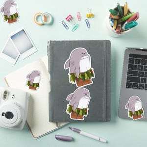 May include: A collection of shark stickers with a cartoon design. The sharks are gray with white bellies, wearing grass skirts and brown boots. The stickers are placed on a notebook, laptop, and other stationery items.