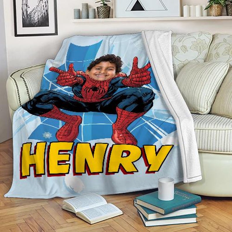 Op de afbeelding: Een lichtblauwe deken met een Spider-Man-ontwerp en een gepersonaliseerde naam in gele blokletters. De deken heeft een stripboekachtige achtergrond en een foto van het gezicht van een persoon in het midden.