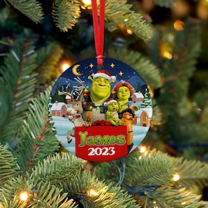 Pode incluir: Um enfeite de Natal redondo com Shrek e outros personagens do filme. O enfeite tem uma fita vermelha para pendurar e inclui o nome "James" e o ano "2023" em uma faixa vermelha.