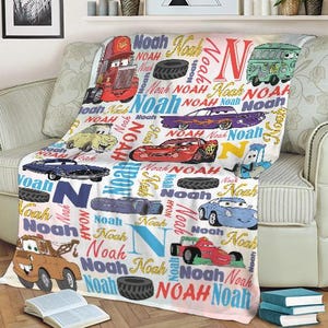 May include: A white blanket featuring various cartoon cars, tires, and the name "Noah" in multiple colors. The blanket is draped over a beige sofa, with a book and stacked books in the foreground.