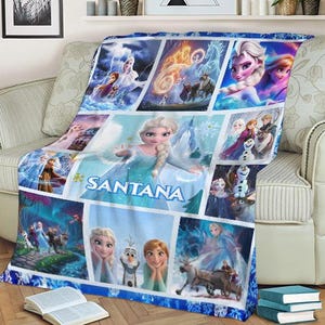 May include: A blue and white fleece blanket featuring a collage of images from the animated film Frozen. The blanket has a personalized name, "SANTANA", and includes images of Elsa, Anna, Olaf, and Kristoff. The blanket is draped over a beige sofa.