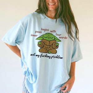 Może przedstawiać: Jasnoniebieski t-shirt z grafiką przedstawiającą zielone stworzenie w brązowej szacie podczas medytacji. Tekst brzmi: inner peace begins with four words: not my fucking problem. Wz&oacute;r jest wyśrodkowany na klatce piersiowej brązowymi i czarnymi literami.