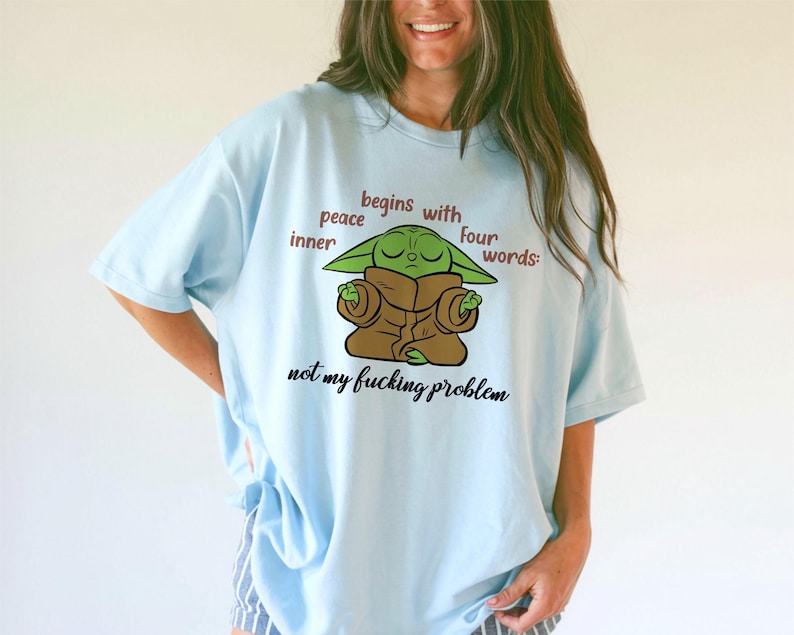 Może przedstawiać: Jasnoniebieski t-shirt z grafiką przedstawiającą zielone stworzenie w brązowej szacie podczas medytacji. Tekst brzmi: inner peace begins with four words: not my fucking problem. Wz&oacute;r jest wyśrodkowany na klatce piersiowej brązowymi i czarnymi literami.